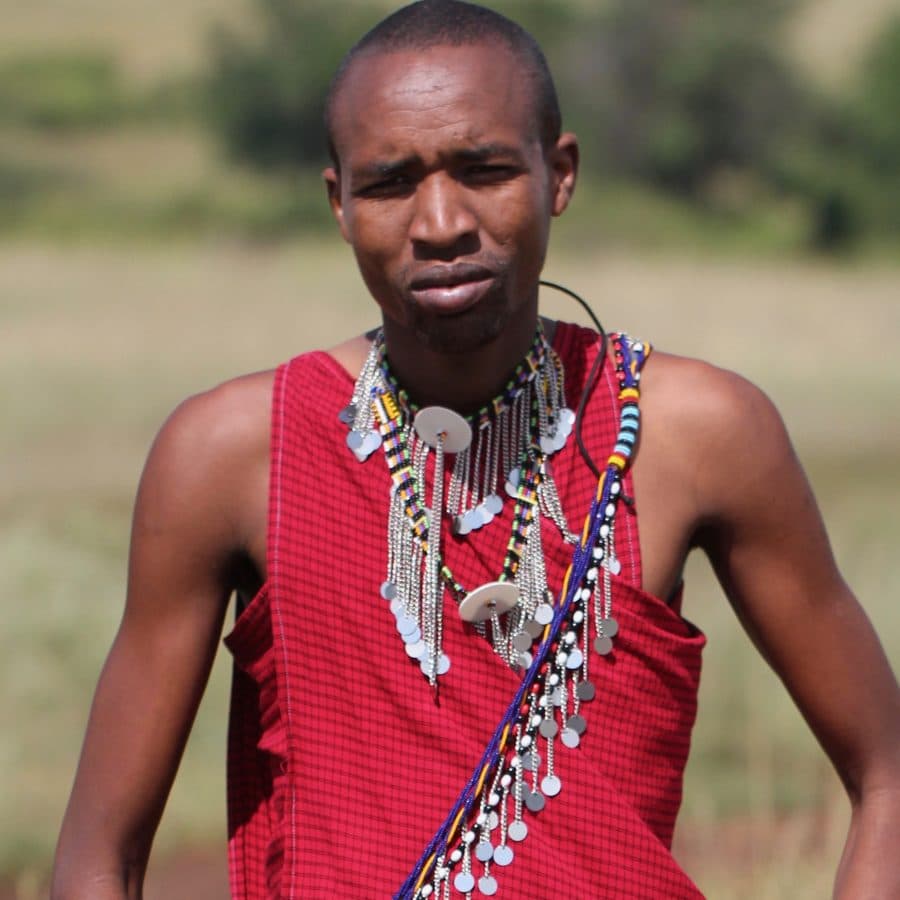 A Maasai man in a red shuka and beaded collar on the open Mara grassland.